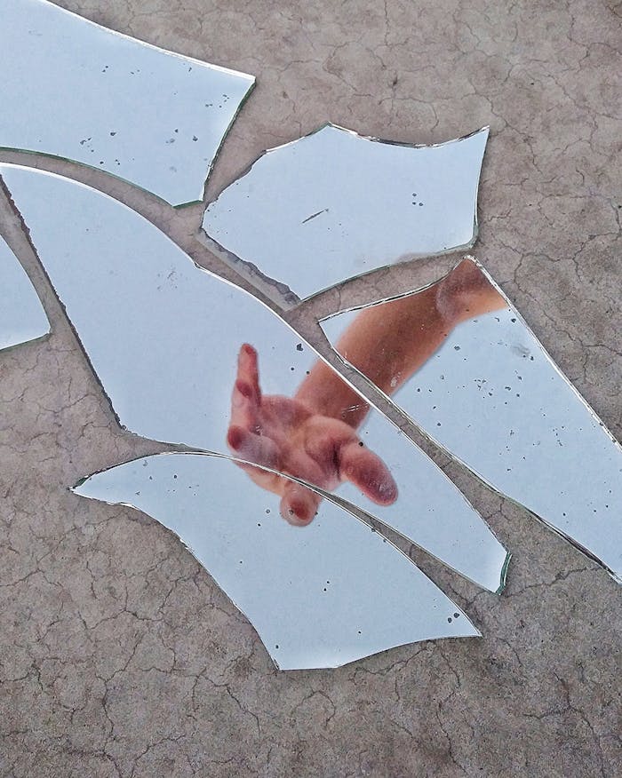 A hand reaching out is reflected in scattered pieces of a broken mirror on a textured surface.